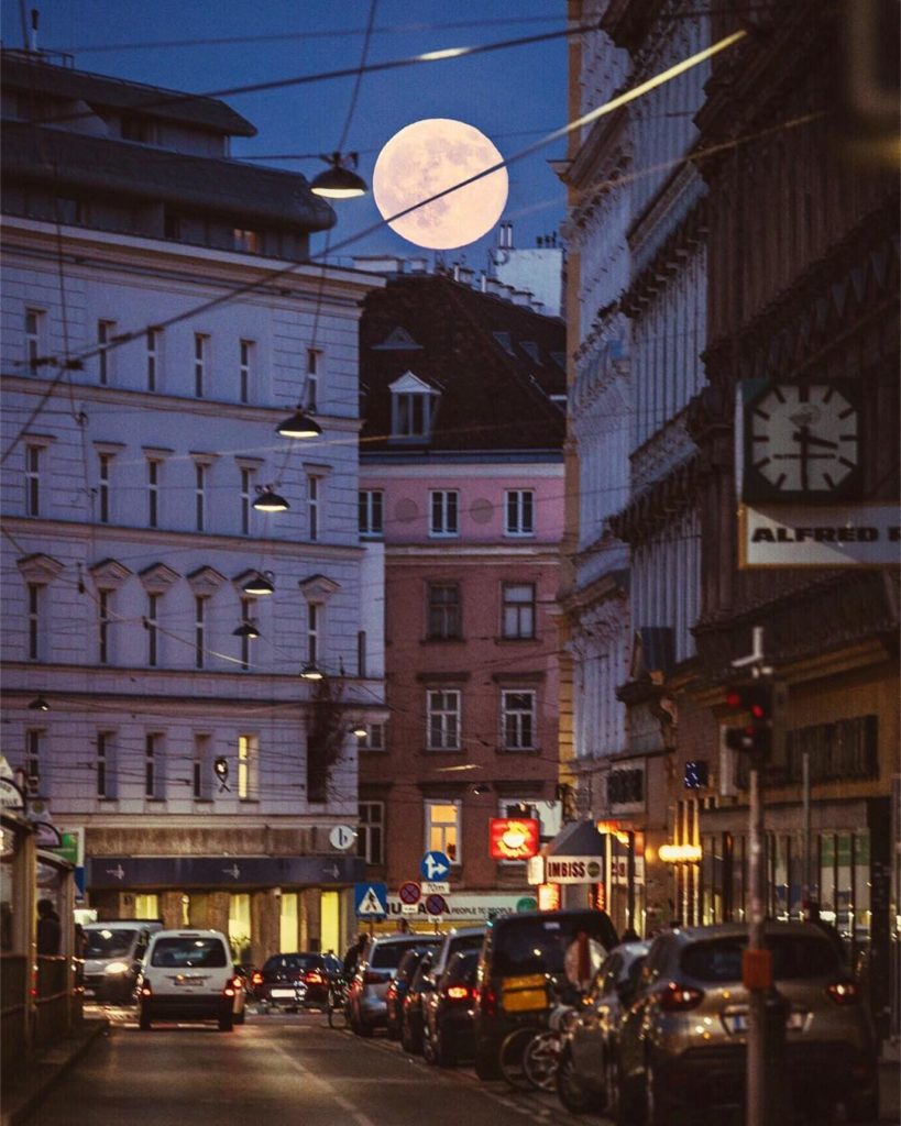  A stunning image captured in Vienna, Austria. The bright supermoon peeps through the
 buildings in a busy street.